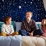 A diverse group of happy children, fully clothed in modest, comfortable pajamas and appropriate attire, enjoying a magical themed pajama party in a beautifully decorated indoor space. The room is transformed into a dreamy night sky with soft, warm fairy lights, projected stars on the ceiling, and cozy piles of soft pillows and blankets creating inviting nooks. Children are casually relaxed, some looking up at the starry projections, others nestled among the cushions. The atmosphere is enchanting and family-friendly. Professional photography, high quality, perfect anatomy, correct proportions, natural pose, well-formed hands, proper finger count, natural body proportions, safe for work, appropriate content, fully clothed.